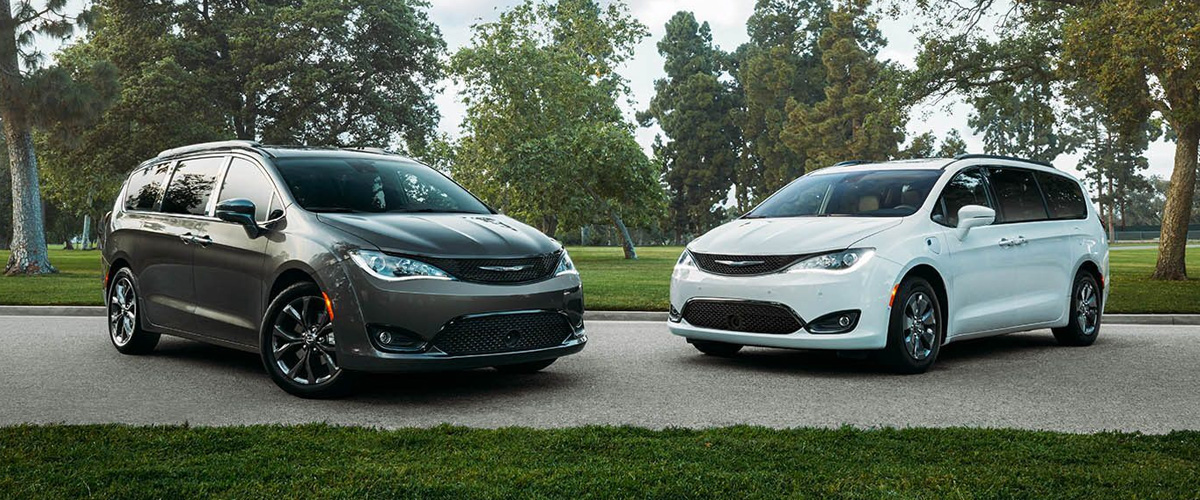 front view of two Chrysler Pacifica vans parked next to each other in front of a park with green trees