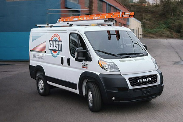 A 2020 Ram ProMaster Cargo Van carrying ladders on its roof rack, being driven in an industrial park.