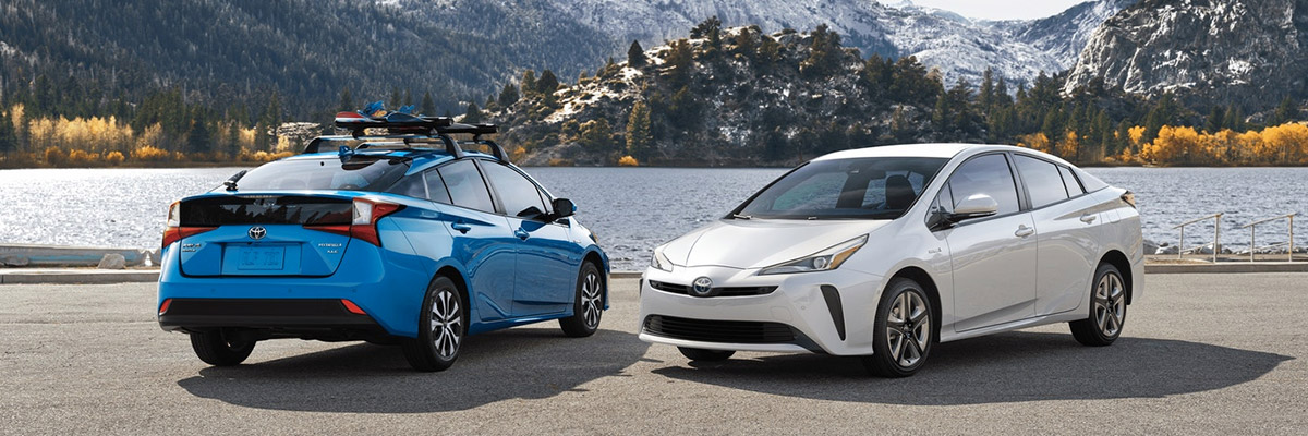 A blue 2020 Toyota Prius parked backwards with a white 2020 Toyota Prius parked forwards right next to it with water and mountains in the background