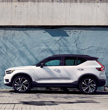 A man stands leaning against a blue concrete facade, next to him is a white Volvo XC40