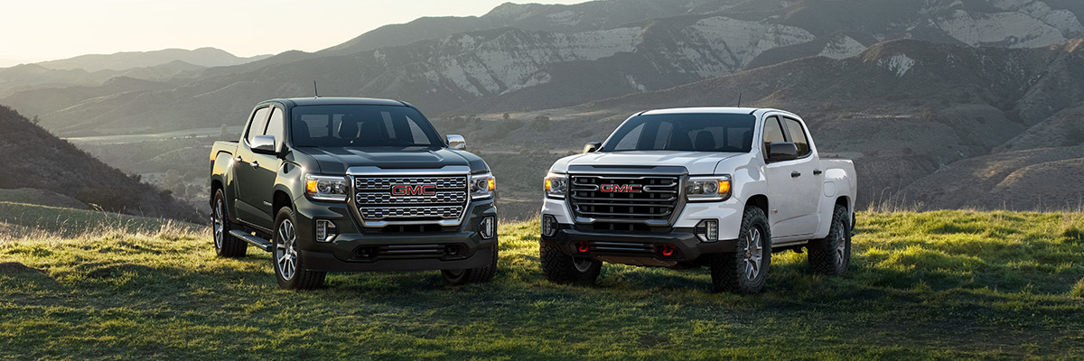2022 GMC Canyon 2-Truck Pack, Family Shot; Featuring (L to R) Canyon Denali Crew Cab Short Box in Hunter Metallic with (RQ9) 20 inch Diamond Cut Machined Aluminum Wheels and Canyon AT4 Crew Cab Short Box in Summit White with (R1U) 17 inch Dark Argent Metallic Cast Aluminum Wheels; Mountains in background.