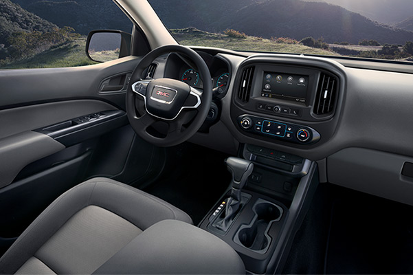2022 GMC Canyon Elevation Standard Interior Cockpit in Jet Black/Dark Ash; Drivers Seat; View from Passengers side rear; Showing Center Console, Cup Holders, Shifter, and Available 7 inch Diagonal Color Touch-Screen Audio System with Navigation and GMC Infotainment; Mountains in background