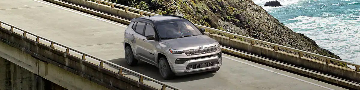 The 2022 Jeep Compass High Altitude being driven over a bridge with a mountain sloping down to the sea in the background.