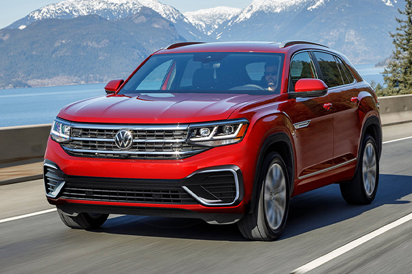 The Atlas Cross Sport in Aurora Red Metallic as seen from the front driving alongside a mountain and lake.