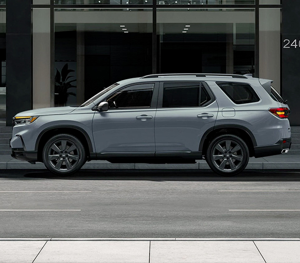Driver-side view of the 2023 Honda Pilot Sport in Sonic Gray Pearl shown parked beside a modern building.