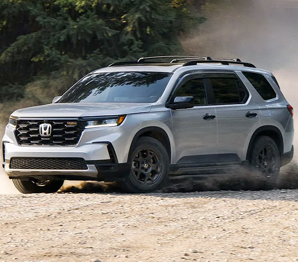 Front driver-side view of the 2023 Honda Pilot TrailSport in Lunar Silver Metallic shown driving on a dirt road through a forest, leaving dust in its wake.