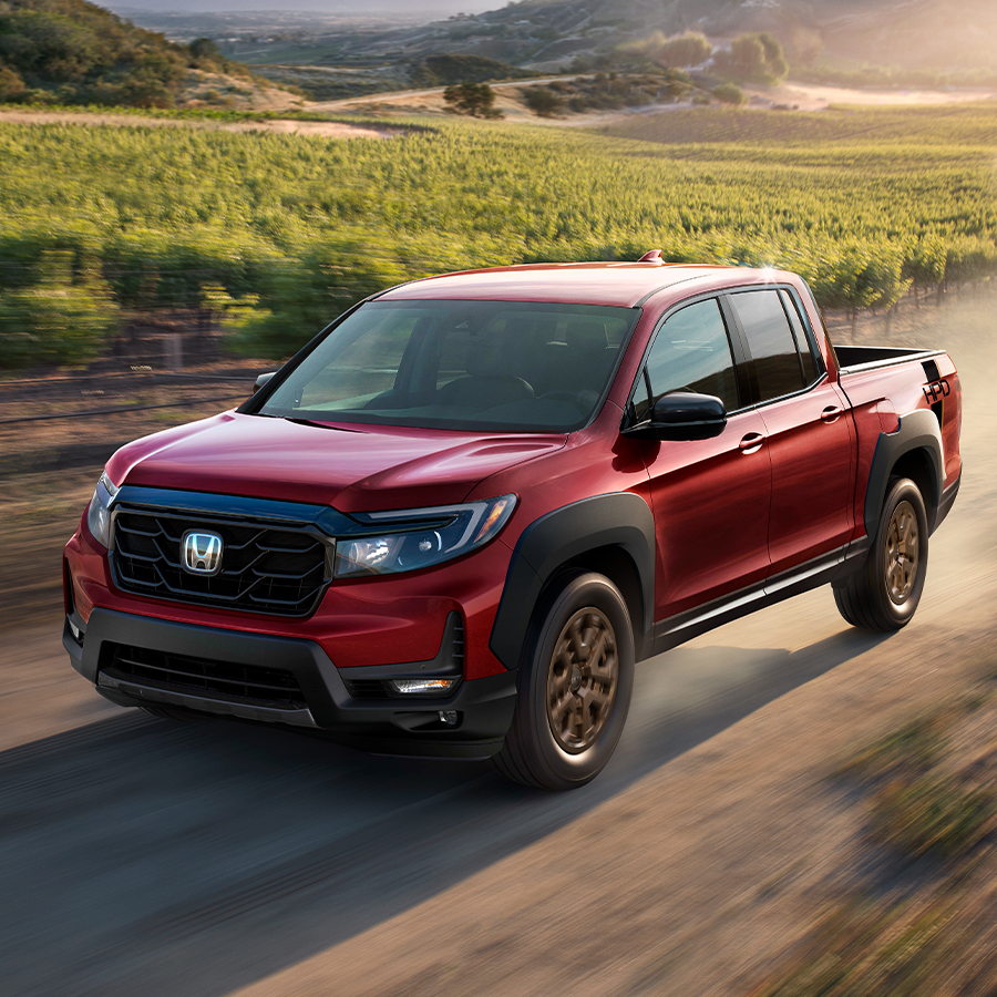 Front driver-side view of the 2023 Honda Ridgeline Sport with available AWD in Radiant Red Metallic II driving down a dusty road.