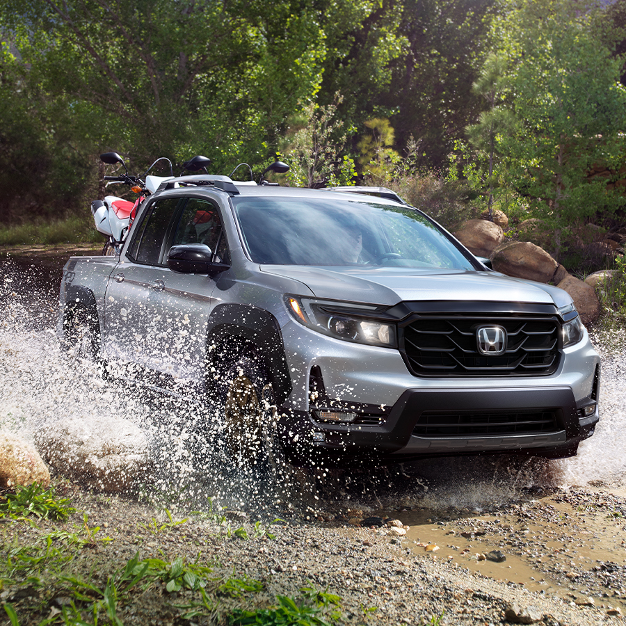 Front passenger-side view of the 2023 Honda Ridgeline Sport in Lunar Silver Metallic with HPD™ Bronze Package, shown driving through a dirt road.
