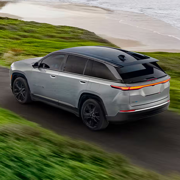 A silver 2024 Jeep Wagoneer S Launch Edition being driven down a beachfront road, with green hills on both sides. The background is blurred to indicate the vehicle is in motion.