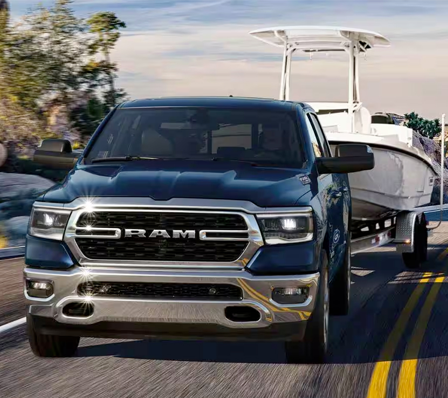 The front end of a blue 2024 Ram 1500 Big Horn Crew Cab towing a speedboat as it is driven down a winding highway in the mountains.
