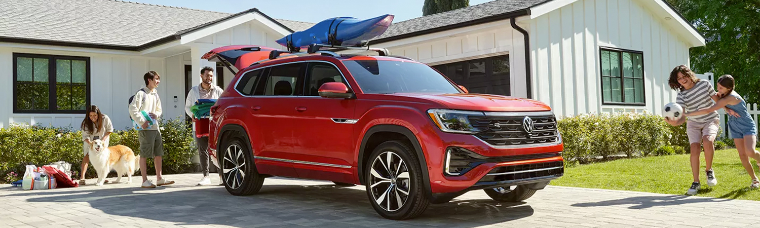 Atlas shown in Aurora Red Metallic parked in the driveway of a mid-century home with a family loading picnic goods in the rear and playing on the lawn nearby.