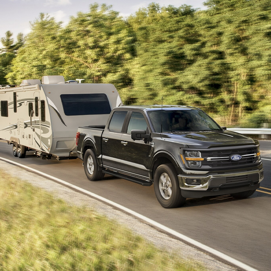 2025 Ford F-150 Platinum in Darkened Bronze Metallic pickup pulling a camper on tree-lined road