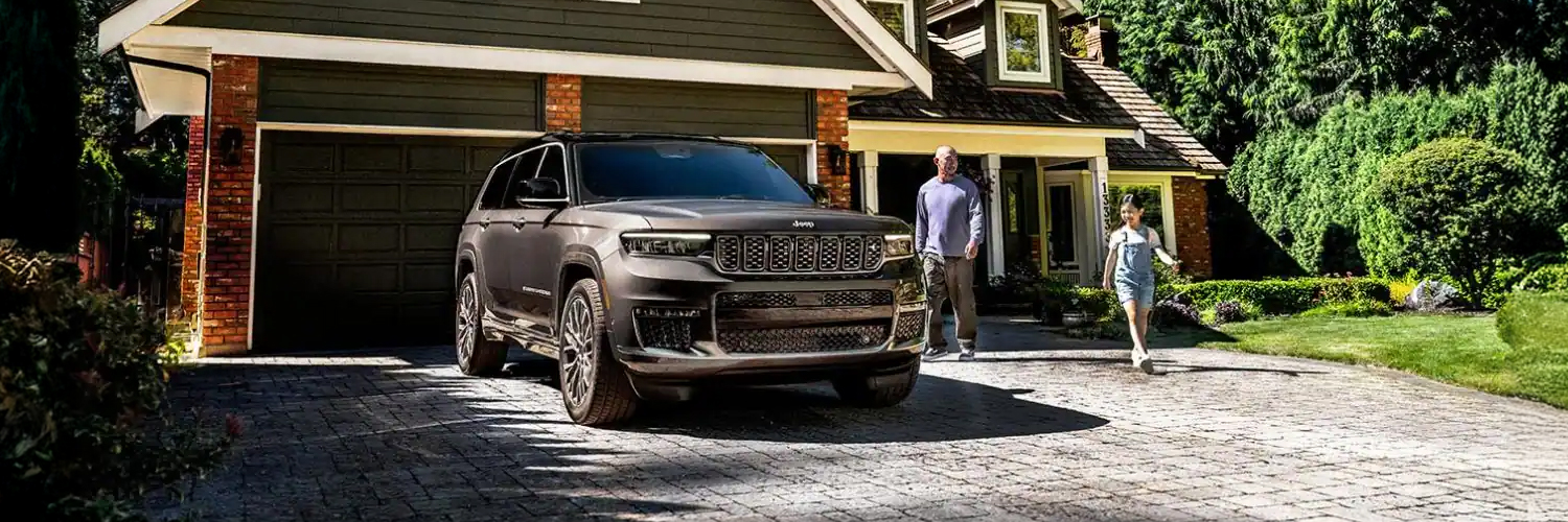 A gray 2025 Jeep Grand Cherokee Summit Reserve parked in the driveway of a neighborhood home with a father and daughter nearby.