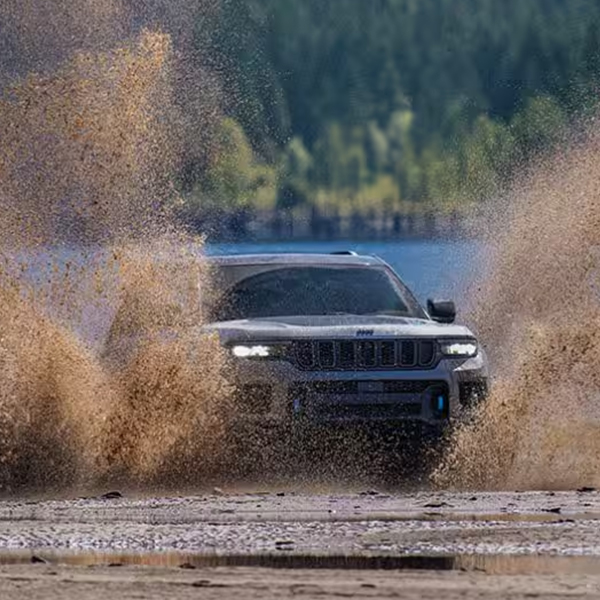 A front angle of a silver 2025 Jeep Grand Cherokee Trailhawk traveling on the muddy shore of a lake with wet sand splashing high above the vehicle on both sides.