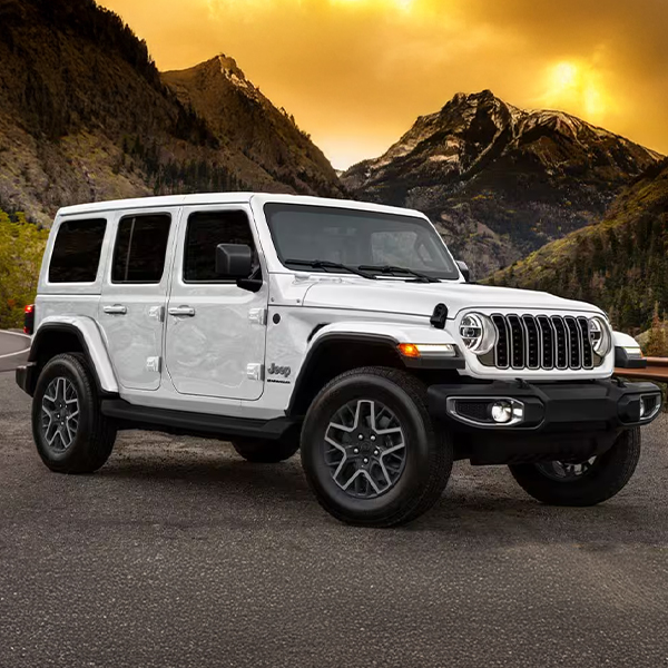 A passenger-side front angle of a white 2025 Jeep Wrangler Sahara parked on the shoulder of a highway in the mountains at sunset.