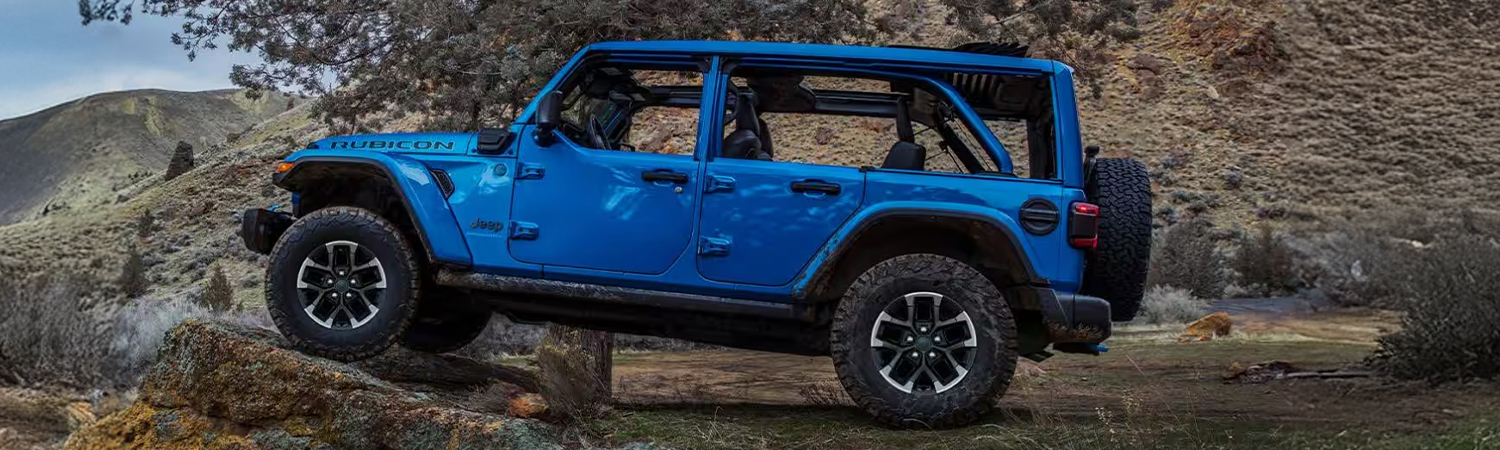 Display A driver-side profile of a blue 2025 Jeep Wrangler Rubicon X 4xe parked off-road, with its front tires elevated on a raised rock formation.