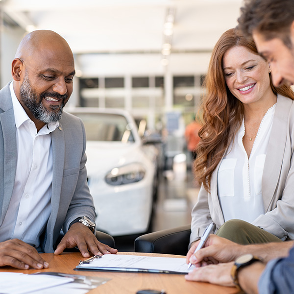 a man purchasing a vehicle and signing a document with a pen while a woman smiles at him